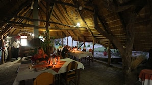 An outdoor dining area with tables and chairs under a thatched roof, surrounded by plants and trees.