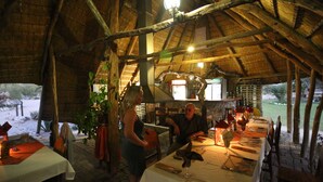 A rustic dining area with wooden beams and a thatched roof. Tables are set with white tablecloths and candles.