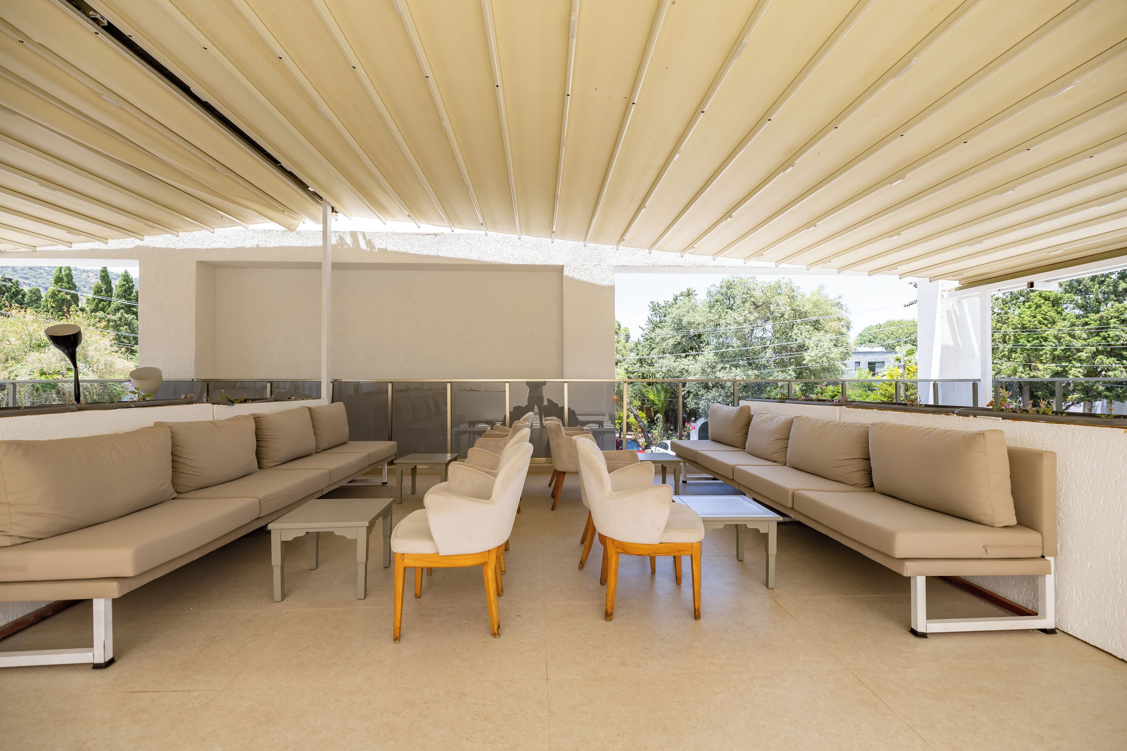 Un espace extérieur aménagé avec des meubles de jardin, comprenant des canapés beiges et des chaises en bois, une table blanche, et offrant une vue sur la verdure et les bâtiments en arrière-plan.