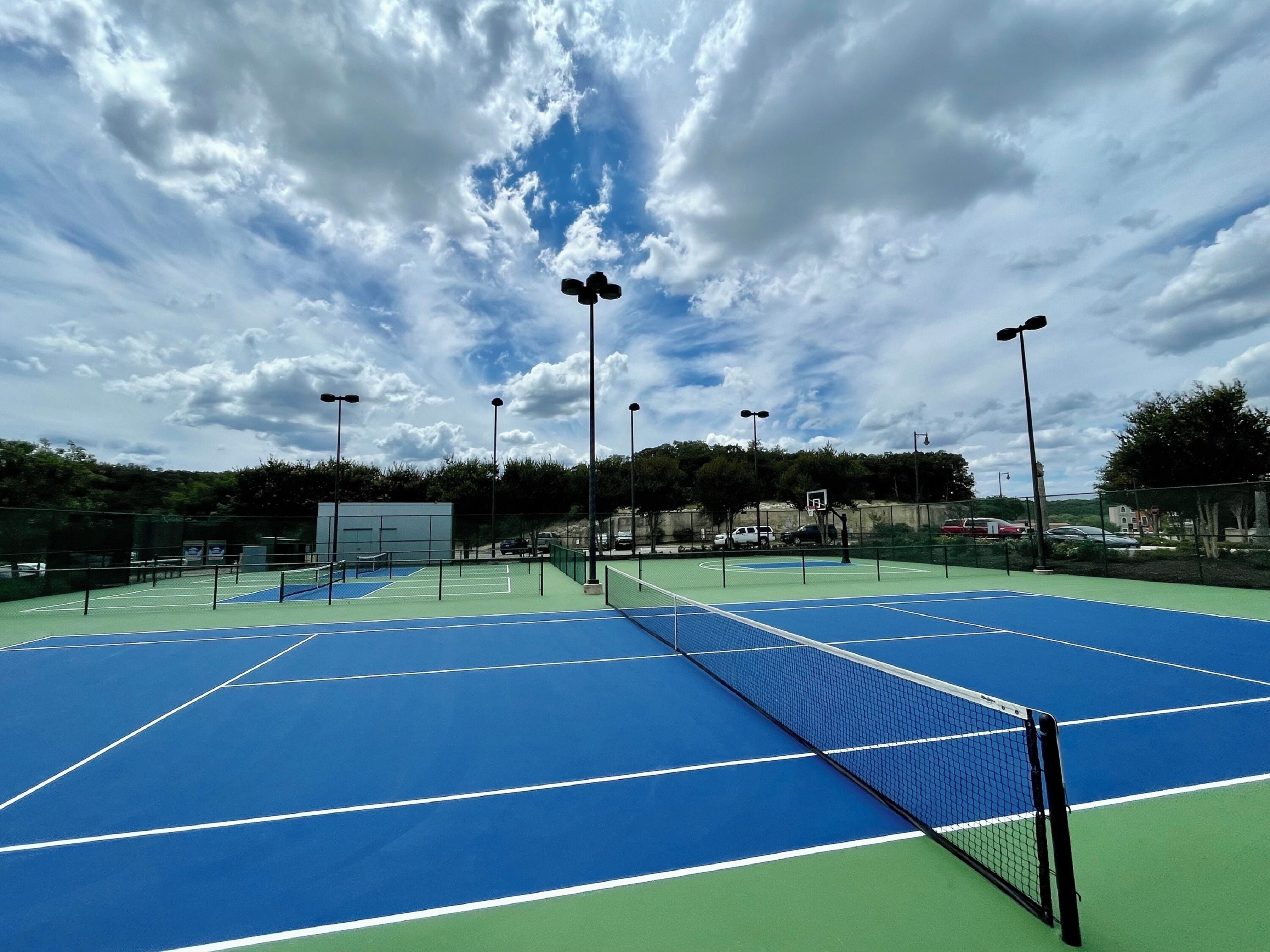 An outdoor tennis court with a net, surrounded by a fence, and under a cloudy sky.