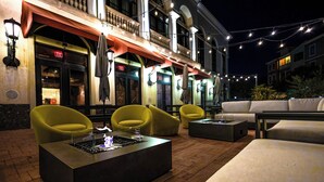 An outdoor seating area with yellow chairs and a fire feature, a building with a red awning, and string lights.