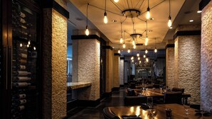 A restaurant interior with a long wooden table, stone walls, hanging light bulbs, and a wine rack.