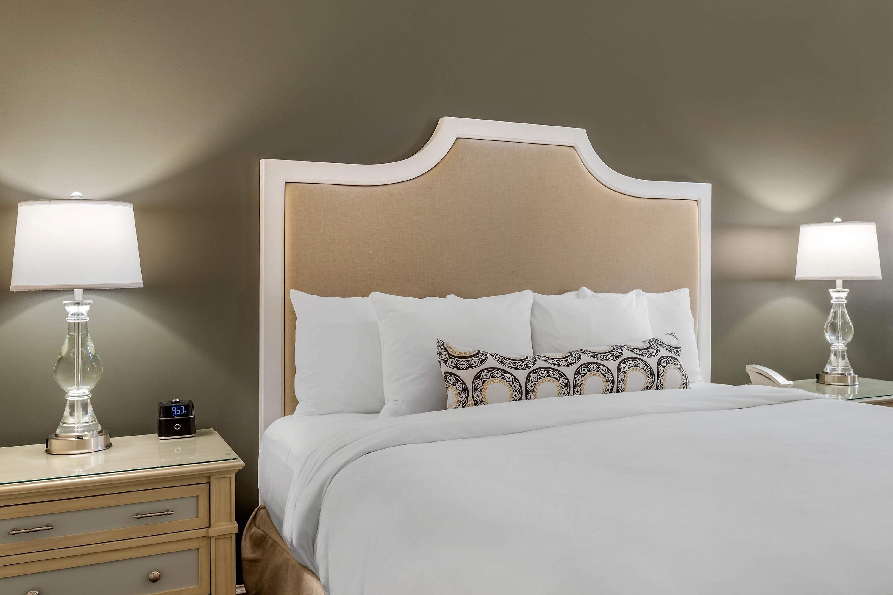 A neatly made bed with white linens and decorative pillows, a bedside table with a lamp, and a nightstand with a clock.