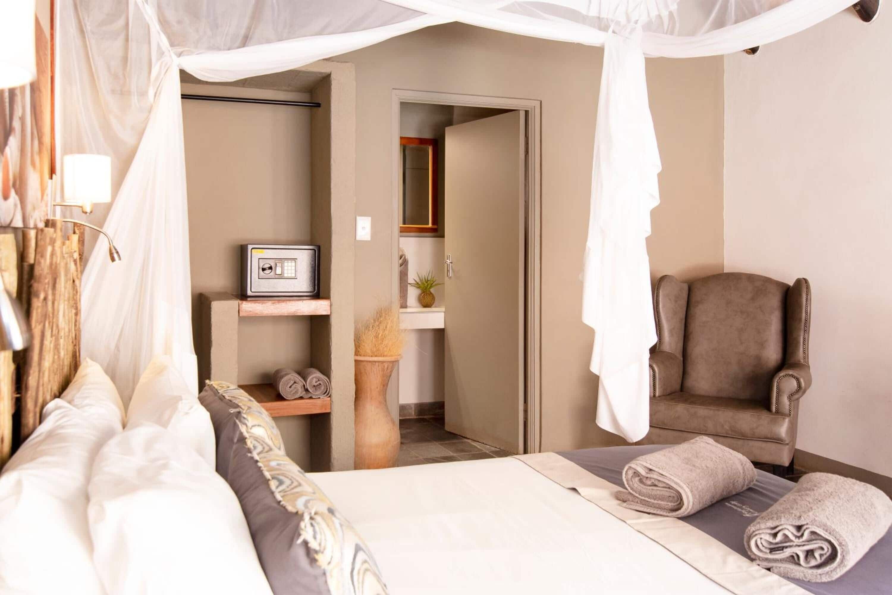 Une chambre à coucher avec un lit à baldaquin, une chaise, une petite table avec une lampe, et une salle de bain visible à travers une porte ouverte.