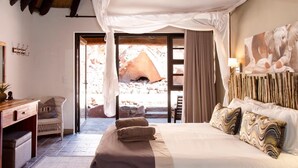 Een slaapkamer met een bed, bureau, stoel en een groot raam met uitzicht op rotsen en een grot.