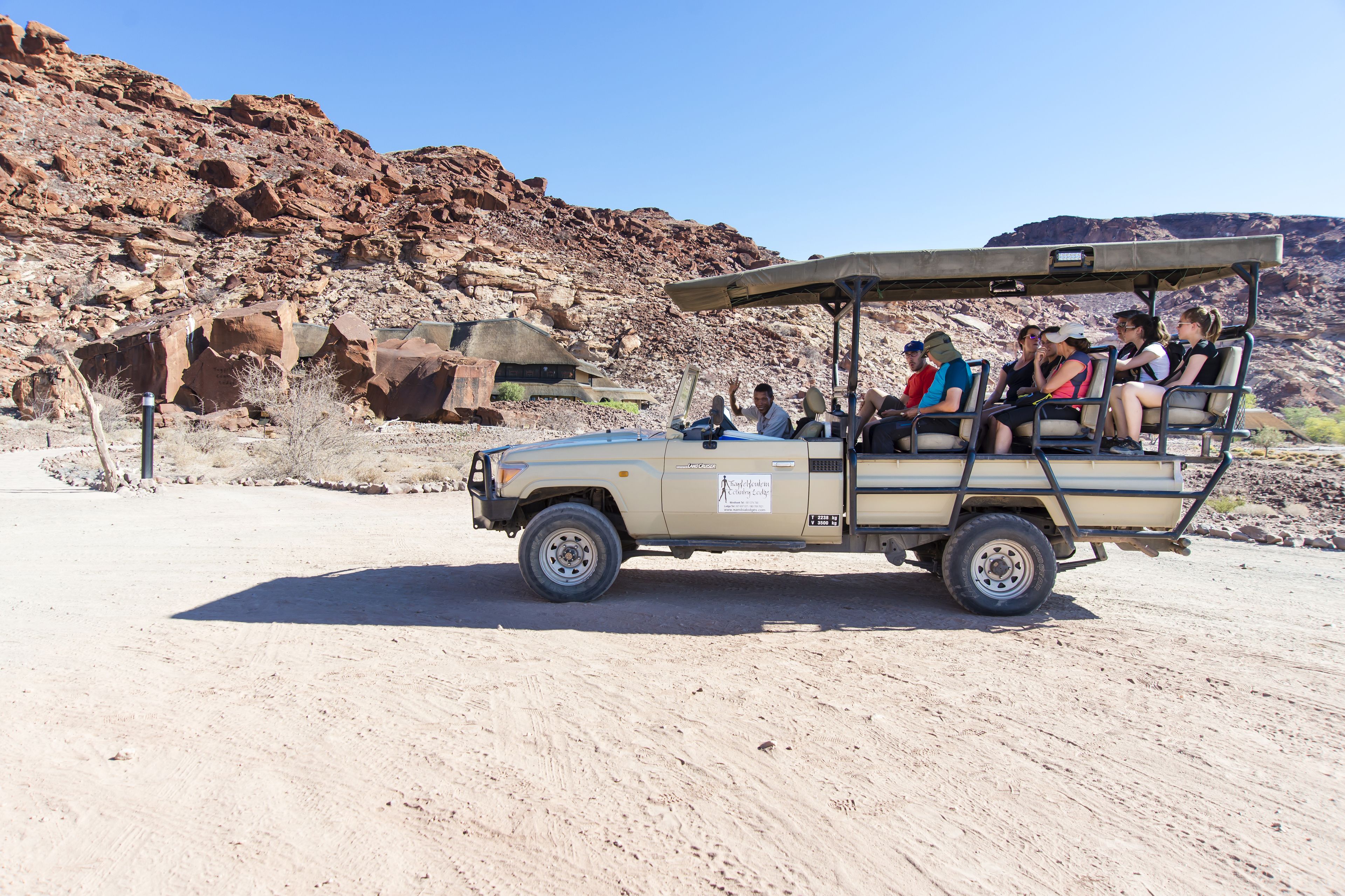 A safari vehicle with passengers, parked in a desert with rocky terrain and sparse vegetation.
