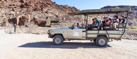 A safari vehicle with passengers, parked in a desert with rocky terrain and sparse vegetation.