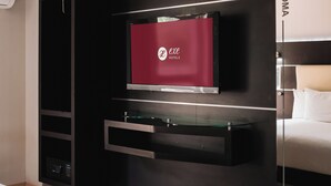 Una habitación de hotel moderna con un televisor de pantalla plana grande montado en una pared negra, una mesa de centro de vidrio y una cama con cabecera que dice "NY.T. ROMA".