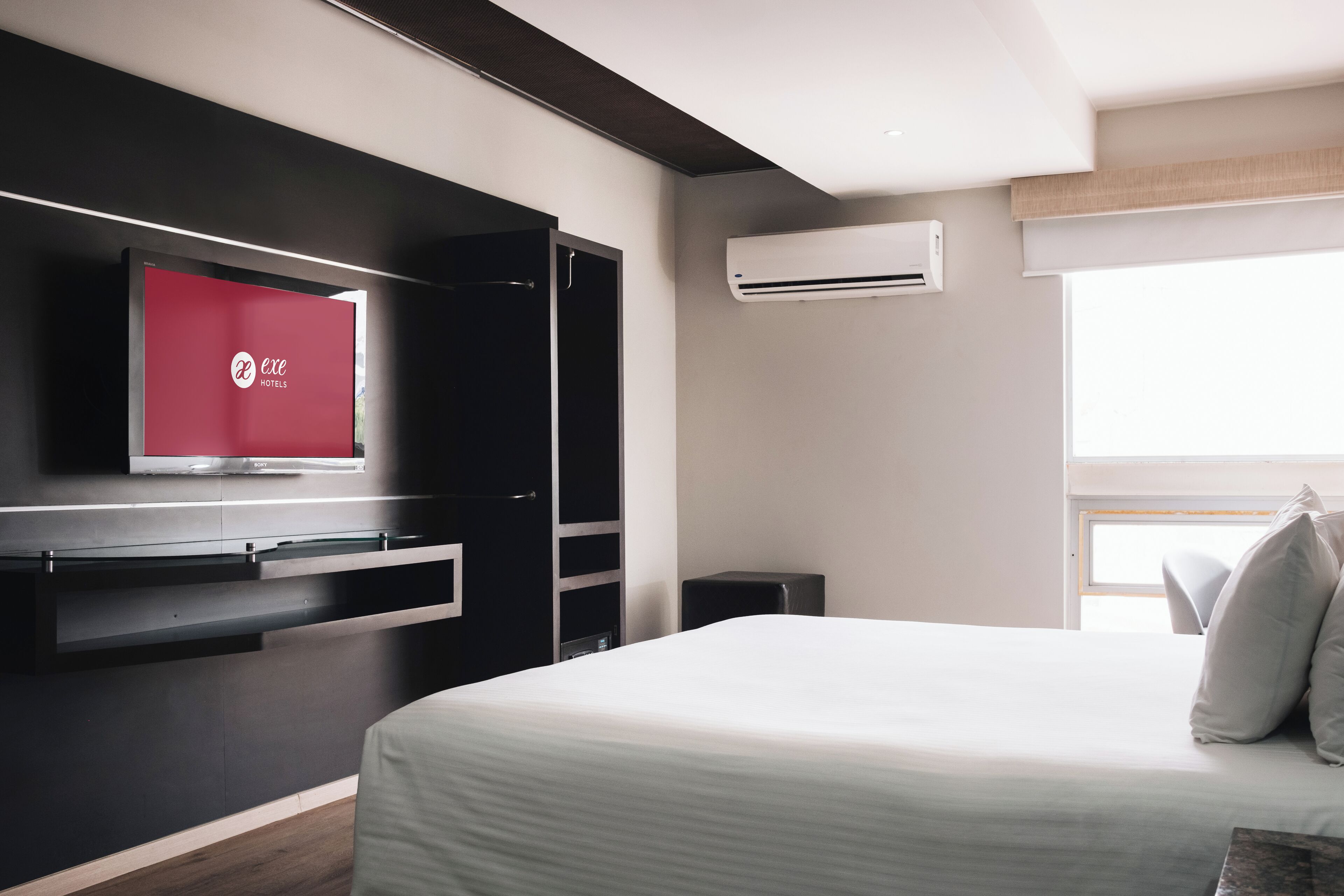 A modern hotel room with a bed, a TV mounted on the wall, a wardrobe, and an air conditioning unit.
