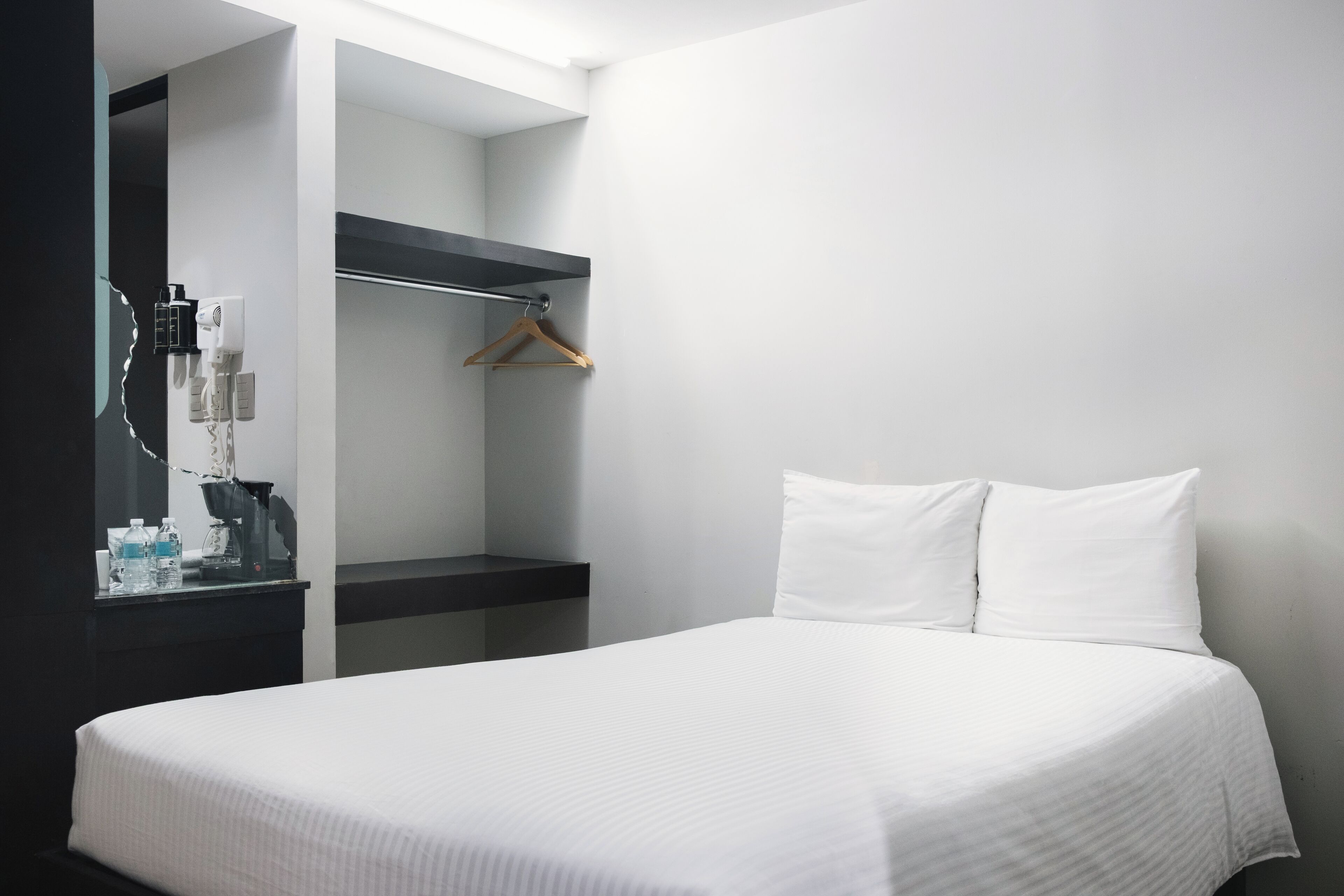 A modern hotel room with a neatly made bed, a black nightstand with a mirror, and a wooden hanger on the wall.
