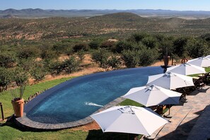 Una piscina de borde curvo, rodeada de sombrillas y sillones blancos, con vistas a un paisaje montañoso.