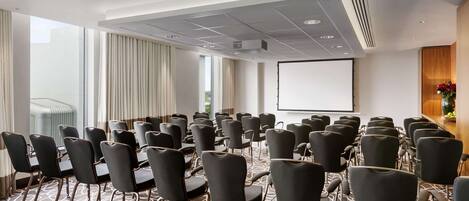 A conference room with rows of chairs, a projector screen, and a large window with curtains.
