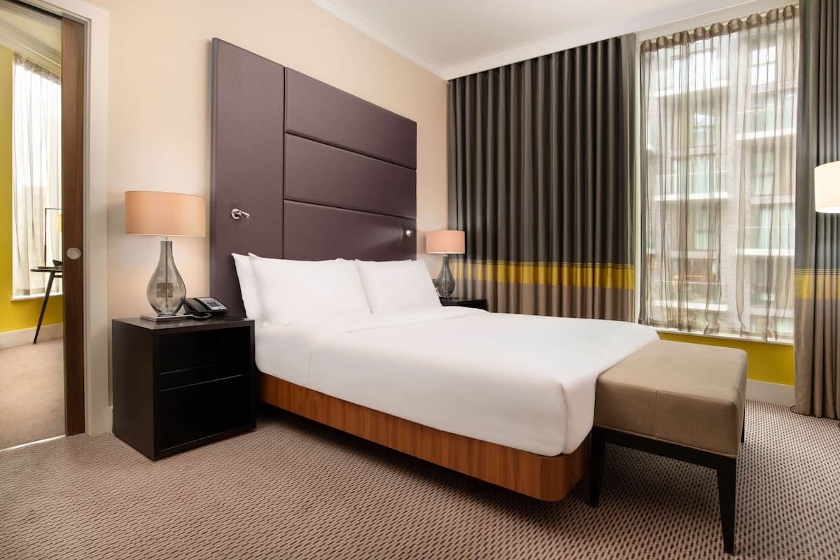 A hotel room with a large bed, two bedside tables with lamps, a bench, and a window with curtains.