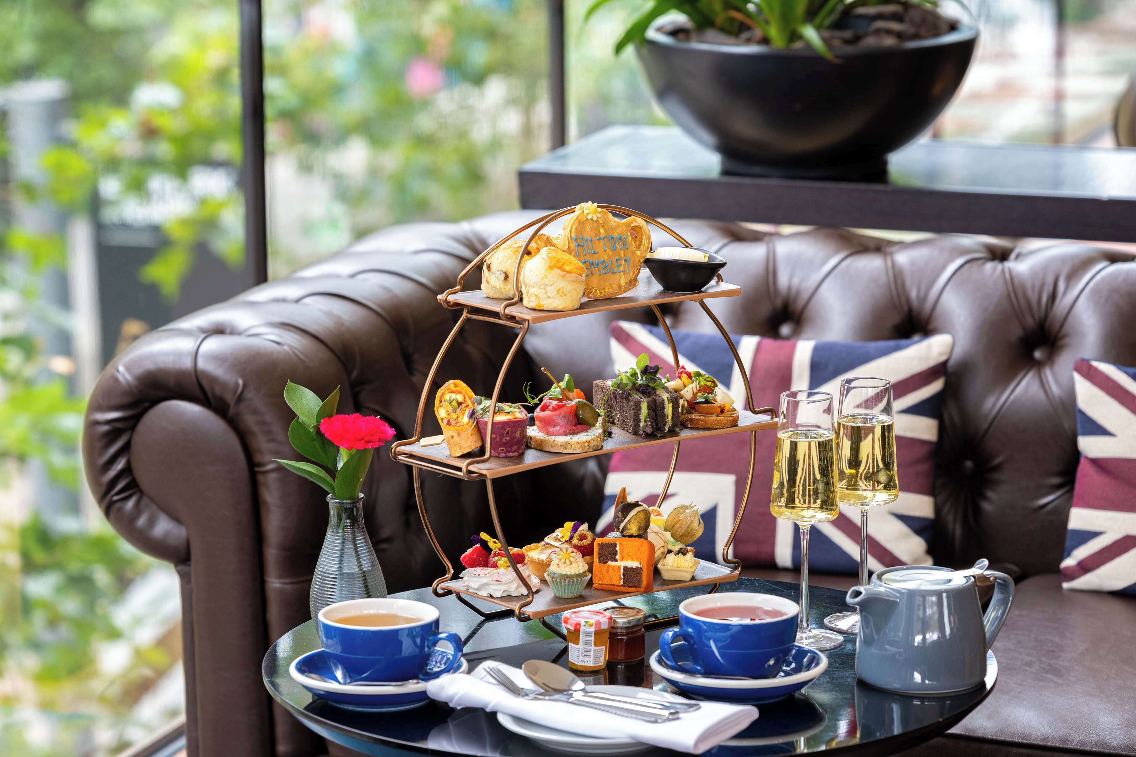 A tiered tray of assorted pastries and sandwiches on a glass table, accompanied by cups of coffee and tea, and a vase with a red flower.