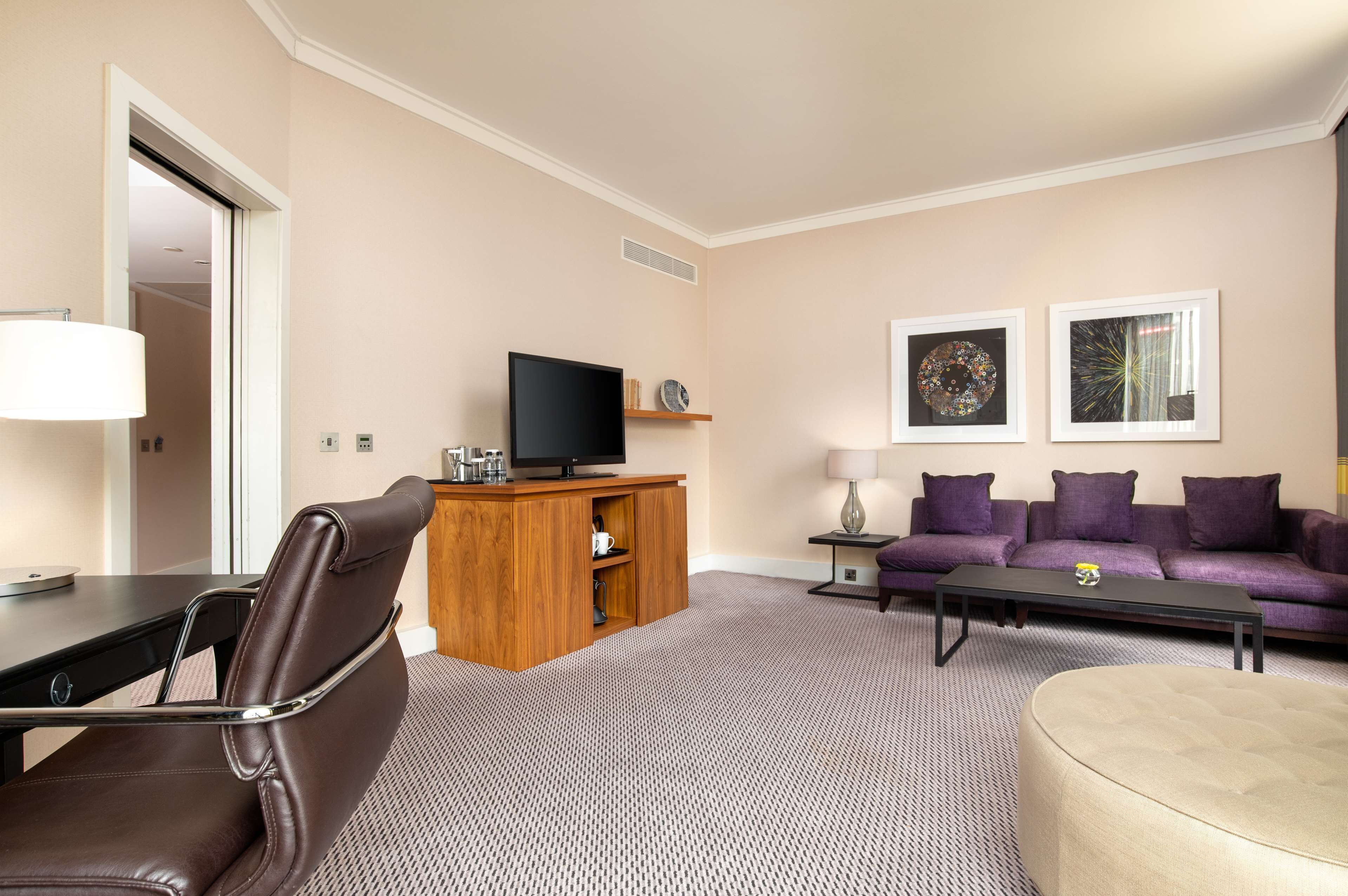 Un salon moderne avec un fauteuil en cuir marron, un meuble de rangement en bois abritant un téléviseur à écran plat, un canapé violet et deux œuvres d’art encadrées accrochées au mur.