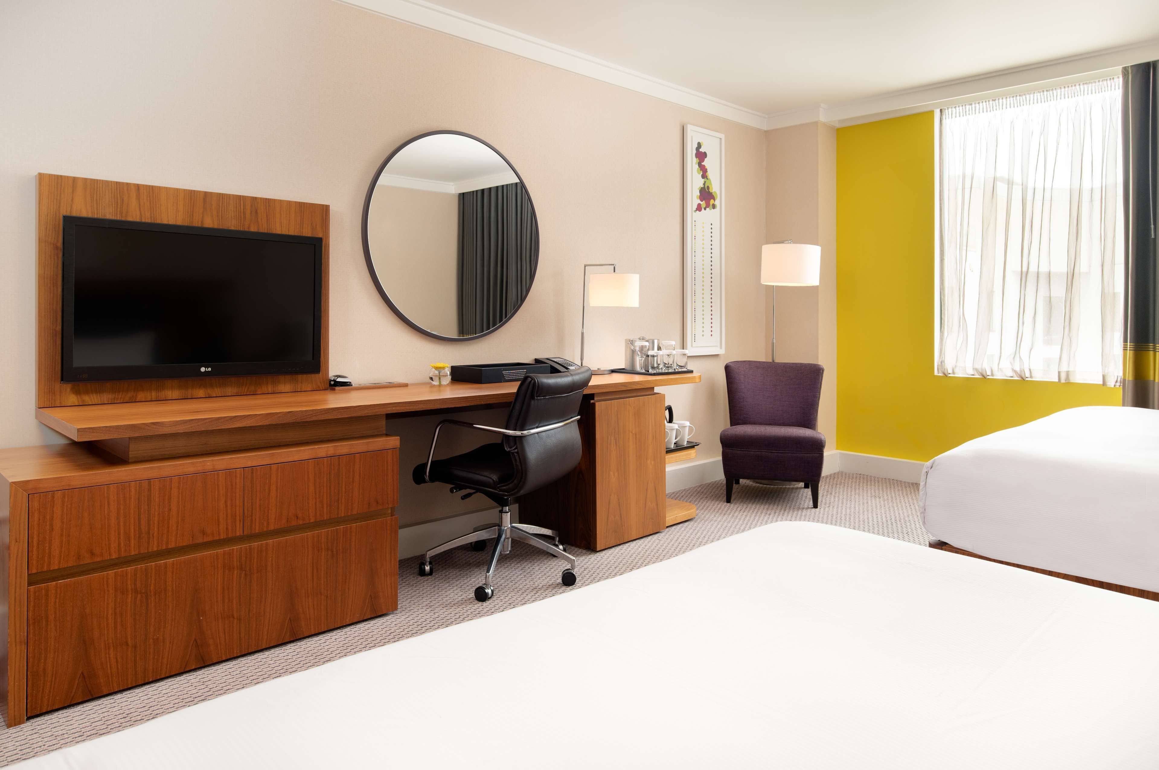 A hotel room with a wooden desk, a flat-screen TV, a round mirror, a purple chair, a bed, and a window with curtains.
