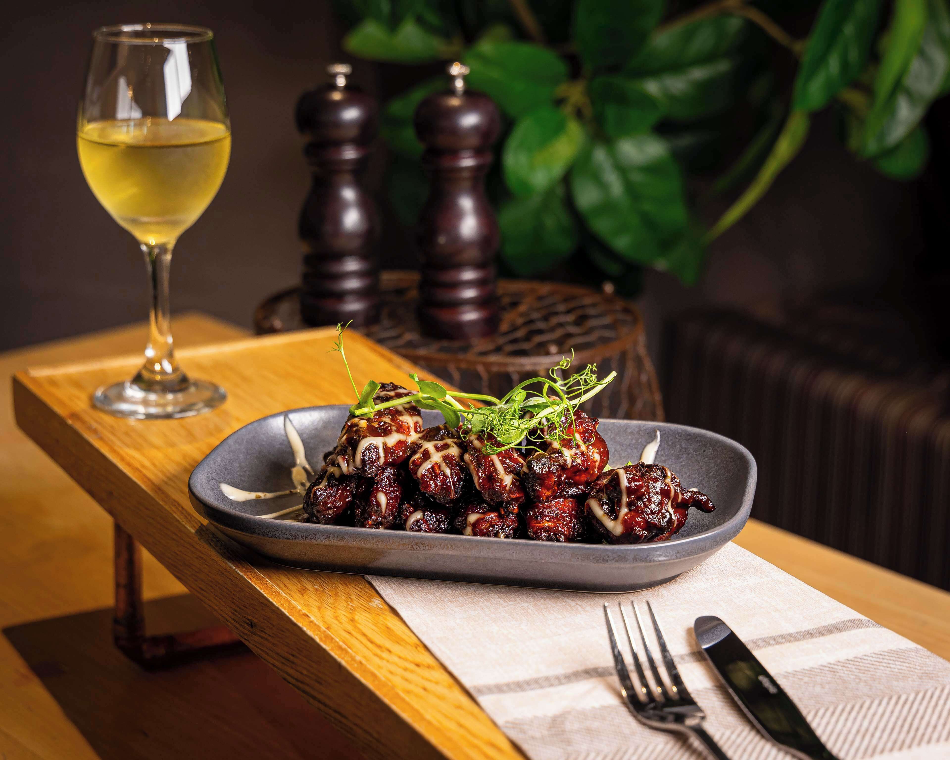A plate of glazed meat garnished with greens, a glass of white wine, and two salt shakers on a wooden table with a napkin and fork.