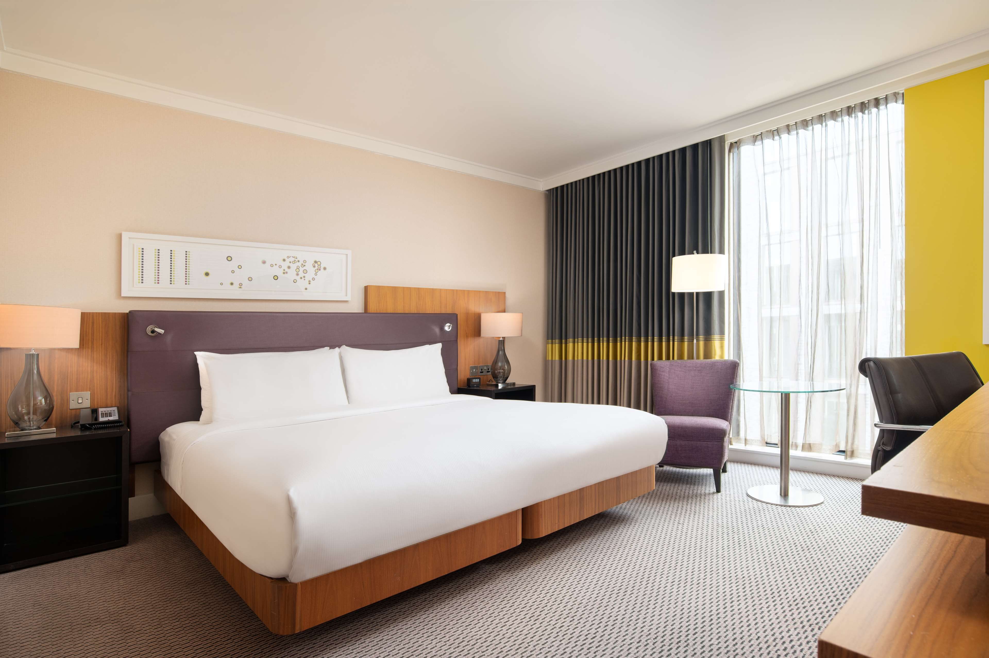 A hotel room with a large bed, a desk, a chair, a lamp, and a view of the city through the window.