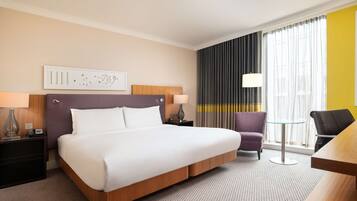A hotel room with a large bed, a desk, a chair, a lamp, and a view of the city through the window.