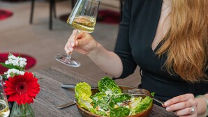 Una persona che tiene un bicchiere di vino bianco, seduta a un tavolo con un'insalata e un vaso di fiori.