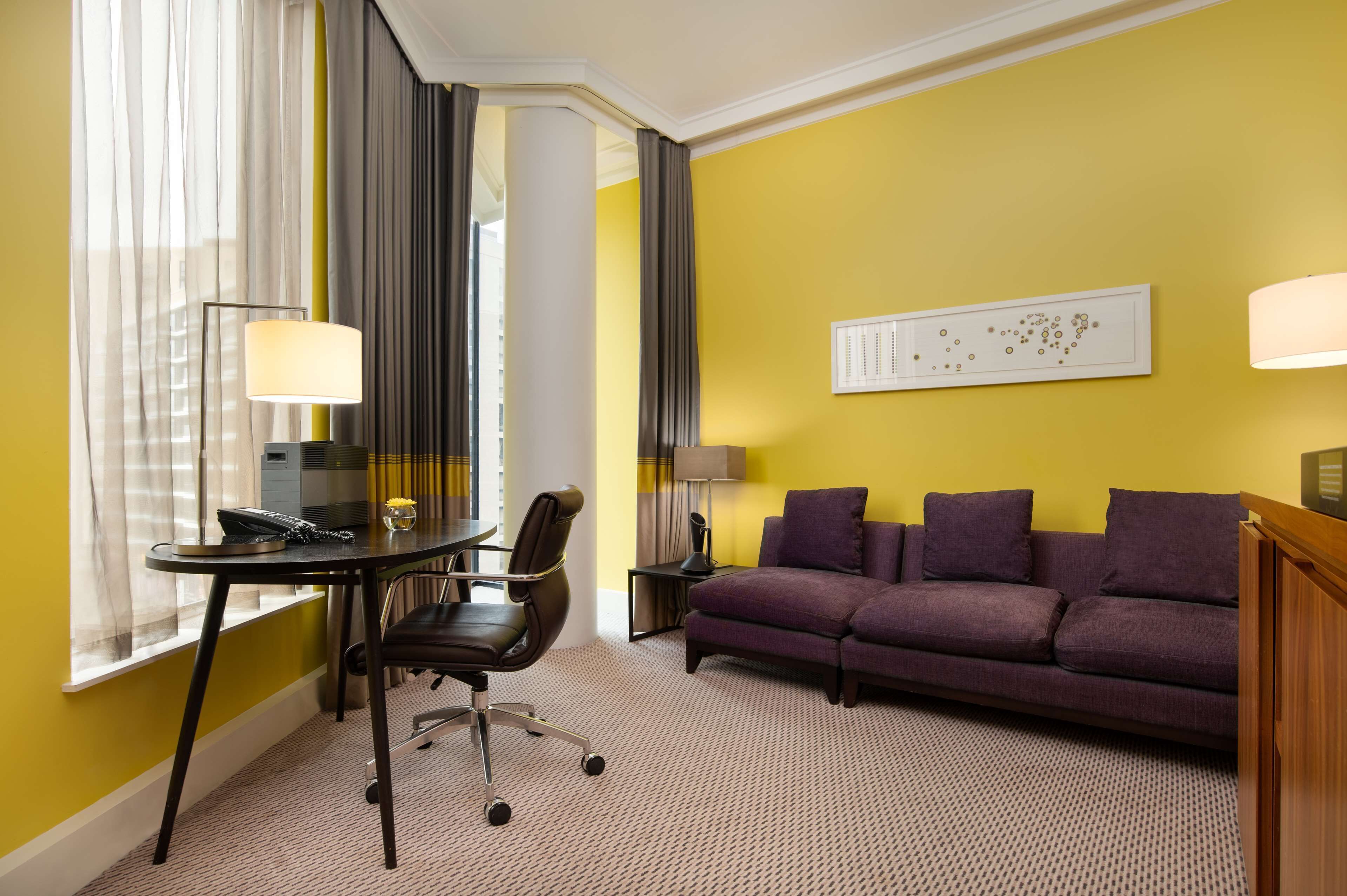 A hotel room with a yellow wall, a purple sofa, a desk with a chair, a lamp, and a window with curtains.