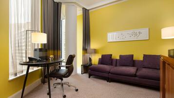 A hotel room with a yellow wall, a purple sofa, a desk with a chair, a lamp, and a window with curtains.