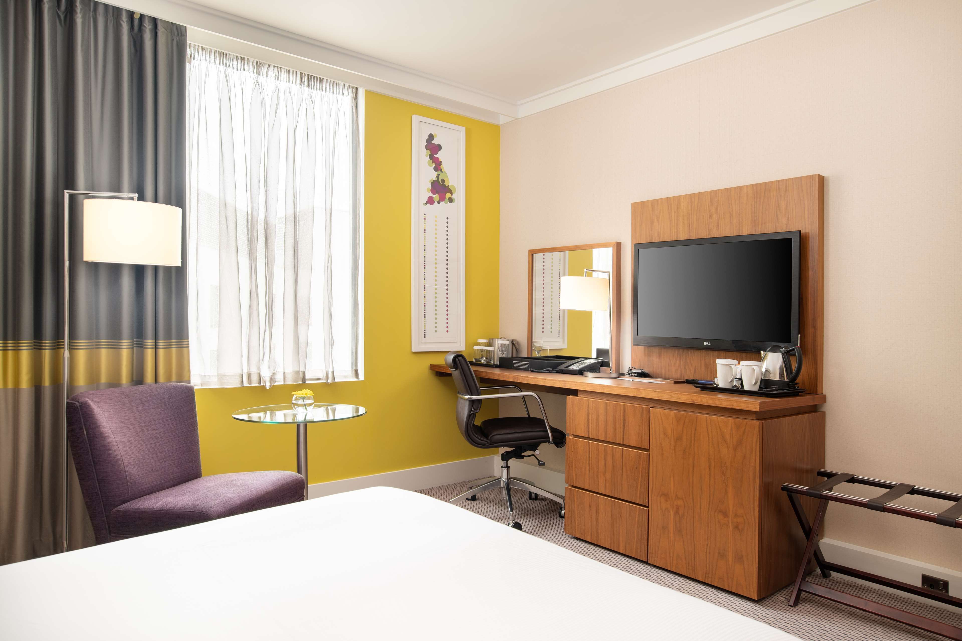 A hotel room with a bed, a desk with a computer, a chair, a small table, a TV, and a window with curtains.