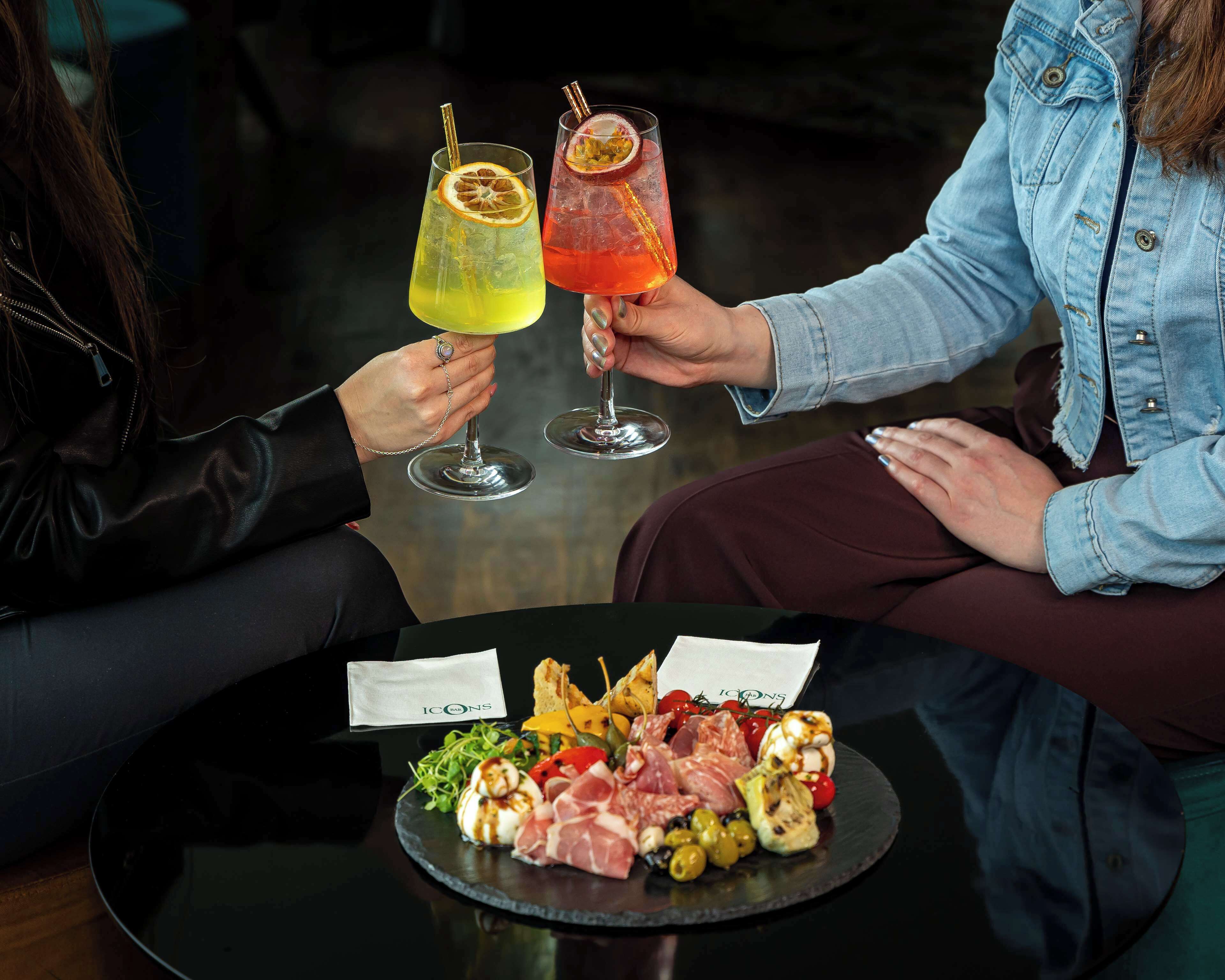 Dos personas brindando con bebidas, un plato de carnes y verduras variadas, y un menú con el logotipo "ICONS".