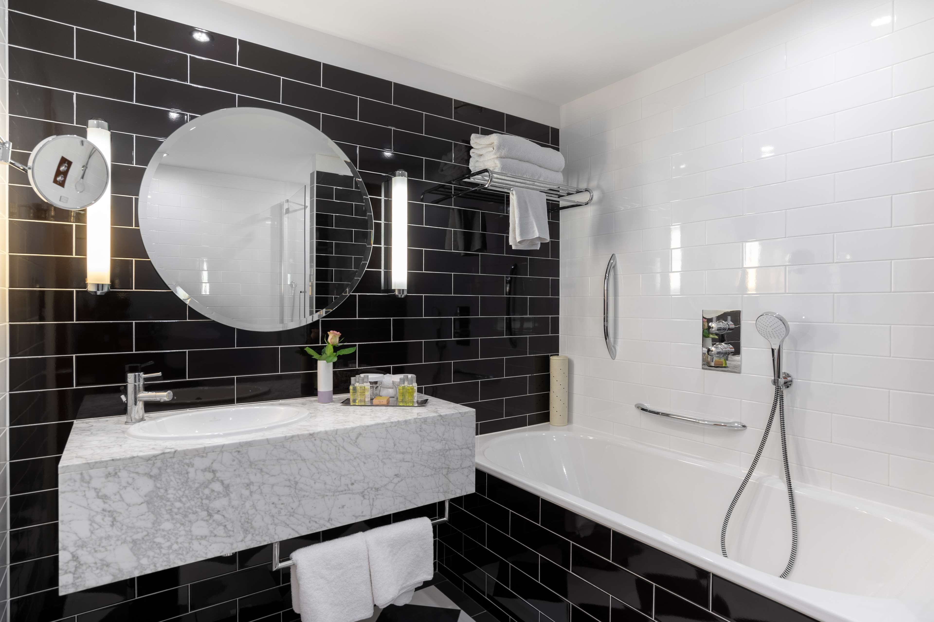 Een moderne badkamer met een witte badkuip, een ronde spiegel, een witte wastafel en zwart betegelde wanden.