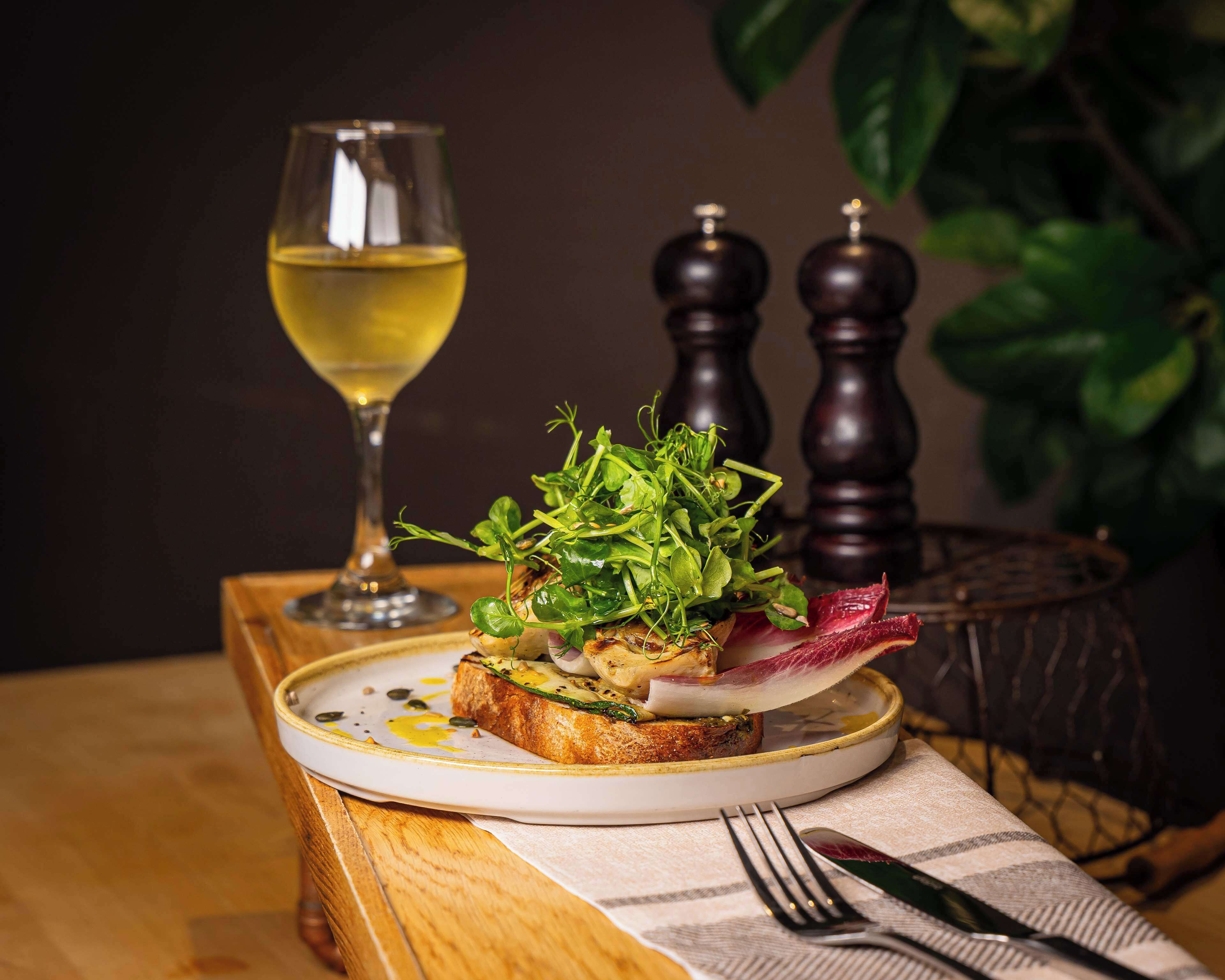 A plate of food with a glass of white wine, a fork, and a knife on a table with a plant and salt and pepper shakers in the background.