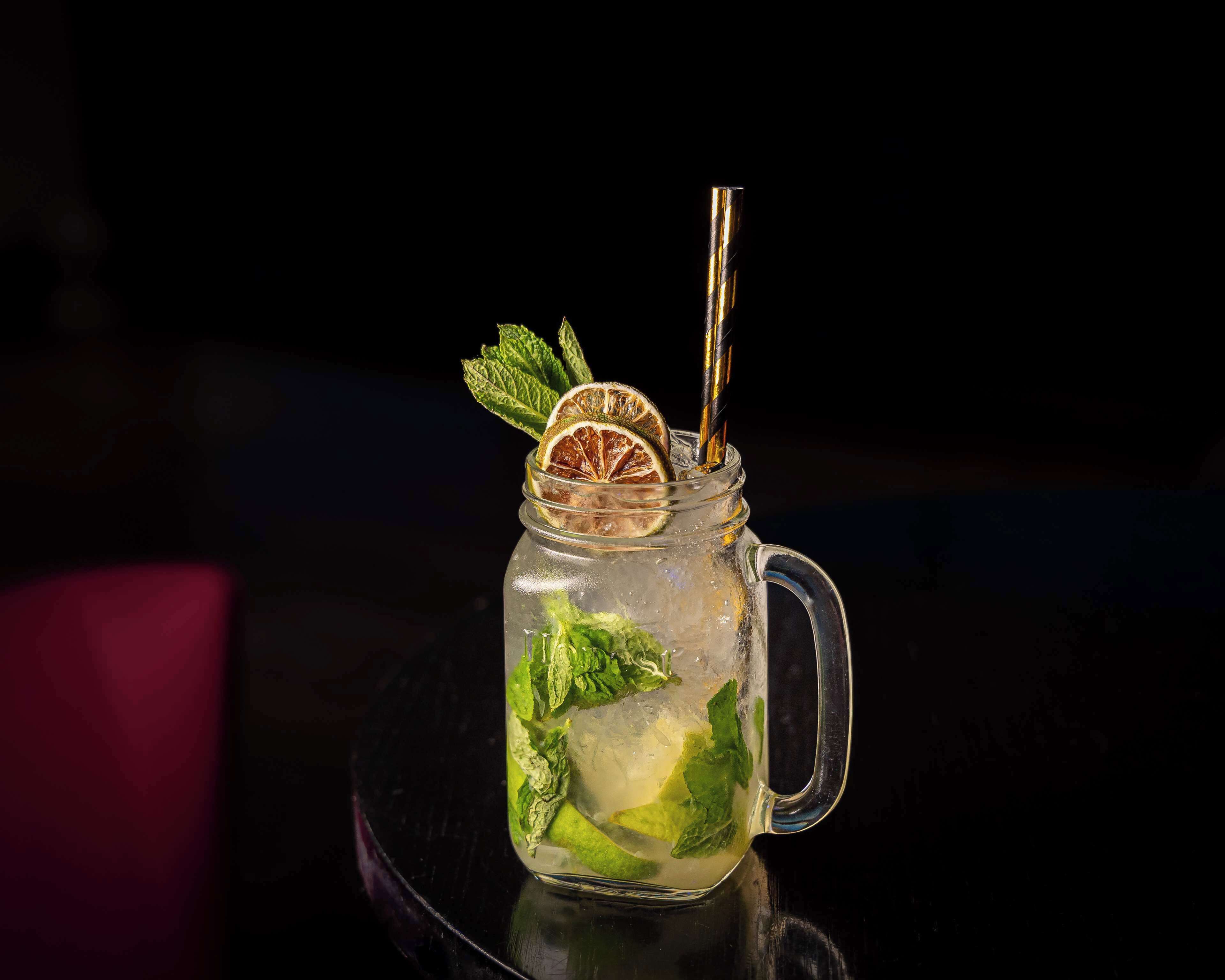 A glass jar with a handle containing a beverage, ice, mint leaves, and a slice of citrus fruit with a straw.