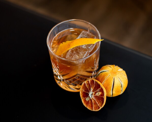 A glass of amber-colored beverage with ice cubes and a slice of dried orange garnish on a dark surface.