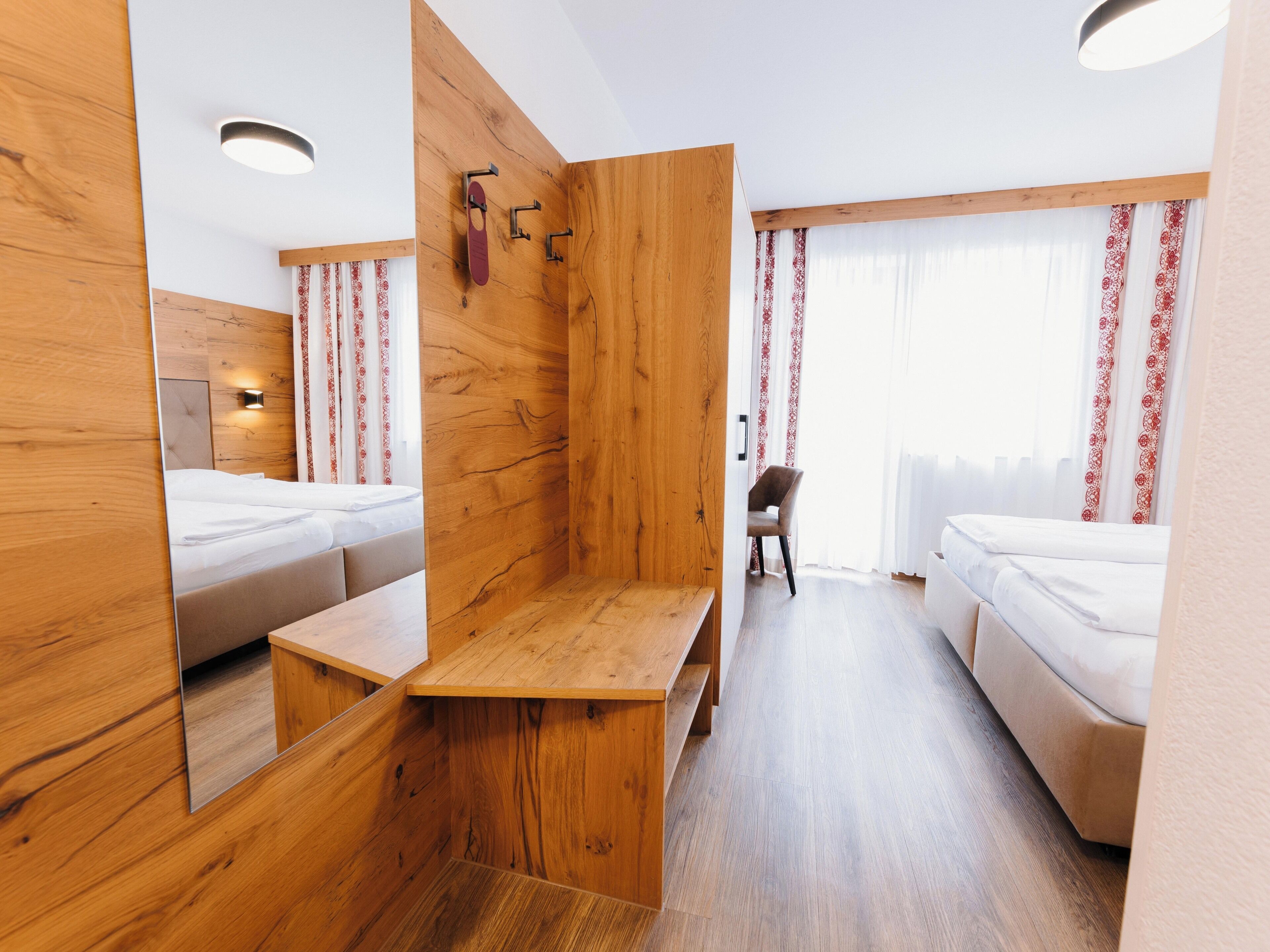 A hotel room with a wooden accent wall, a wooden bench, two beds, a chair, and a mirror.