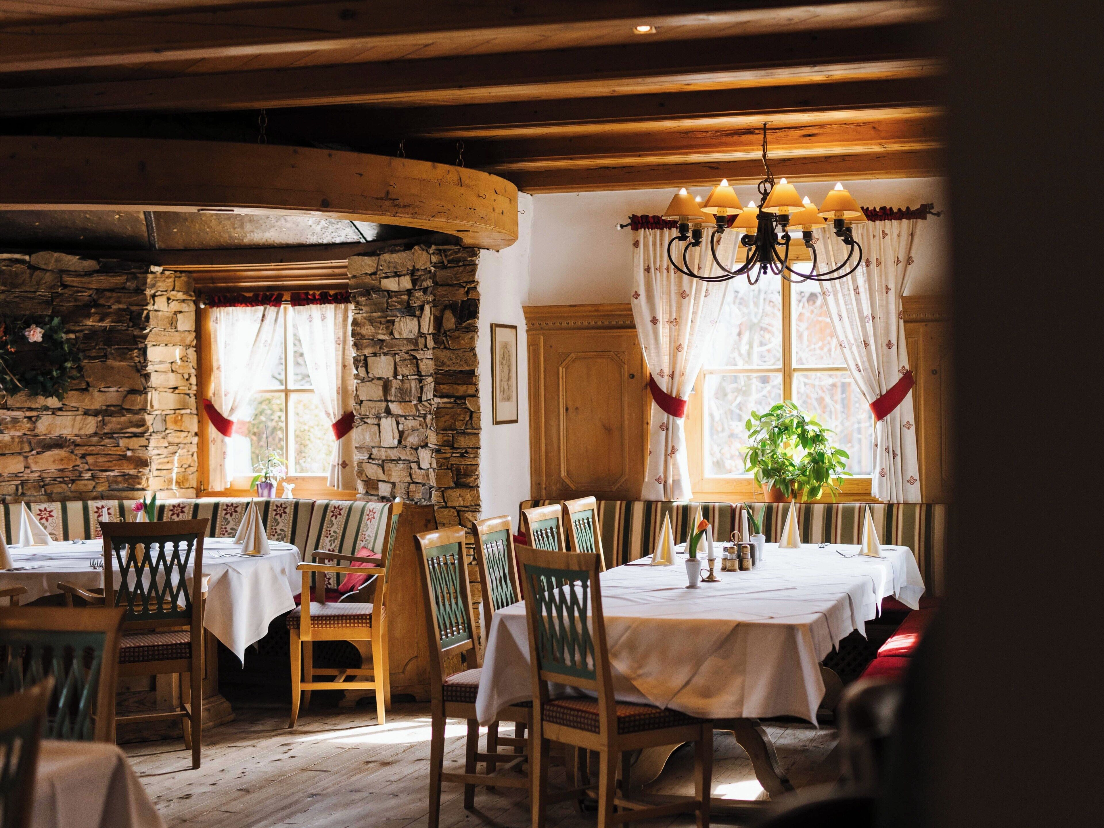 A rustic dining area with wooden beams, stone walls, and a chandelier. Tables are set with white tablecloths and chairs.