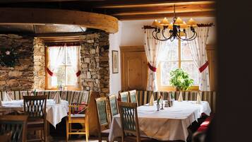 Un coin repas rustique avec des poutres en bois, des murs en pierre et un lustre. Les tables sont dressées avec des nappes blanches et des chaises.