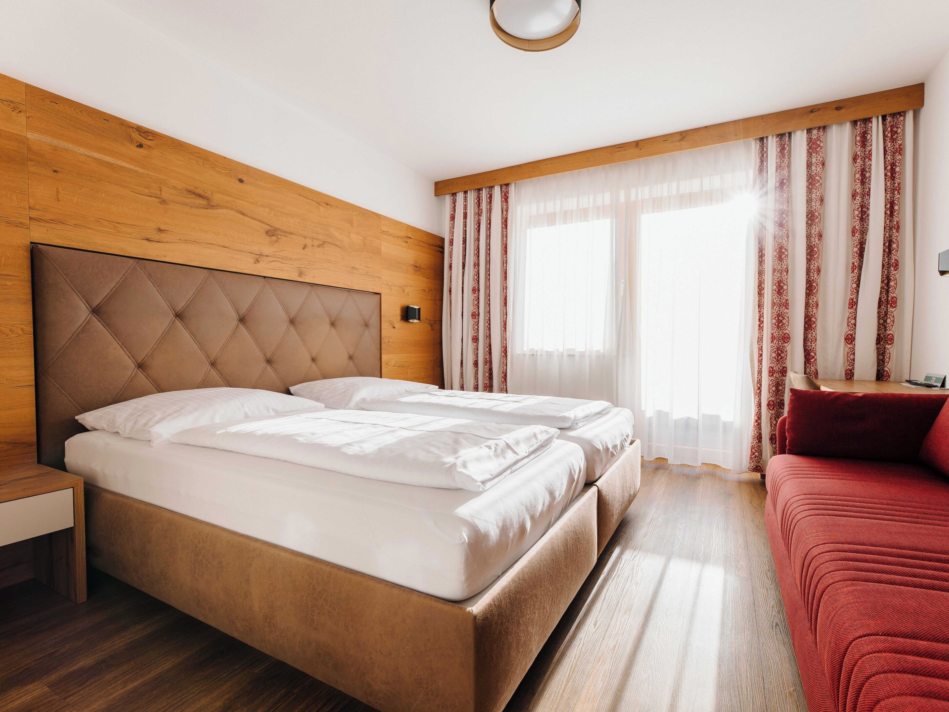 A hotel room with a wooden headboard, a bed with white linens, a red sofa, and a wooden floor.