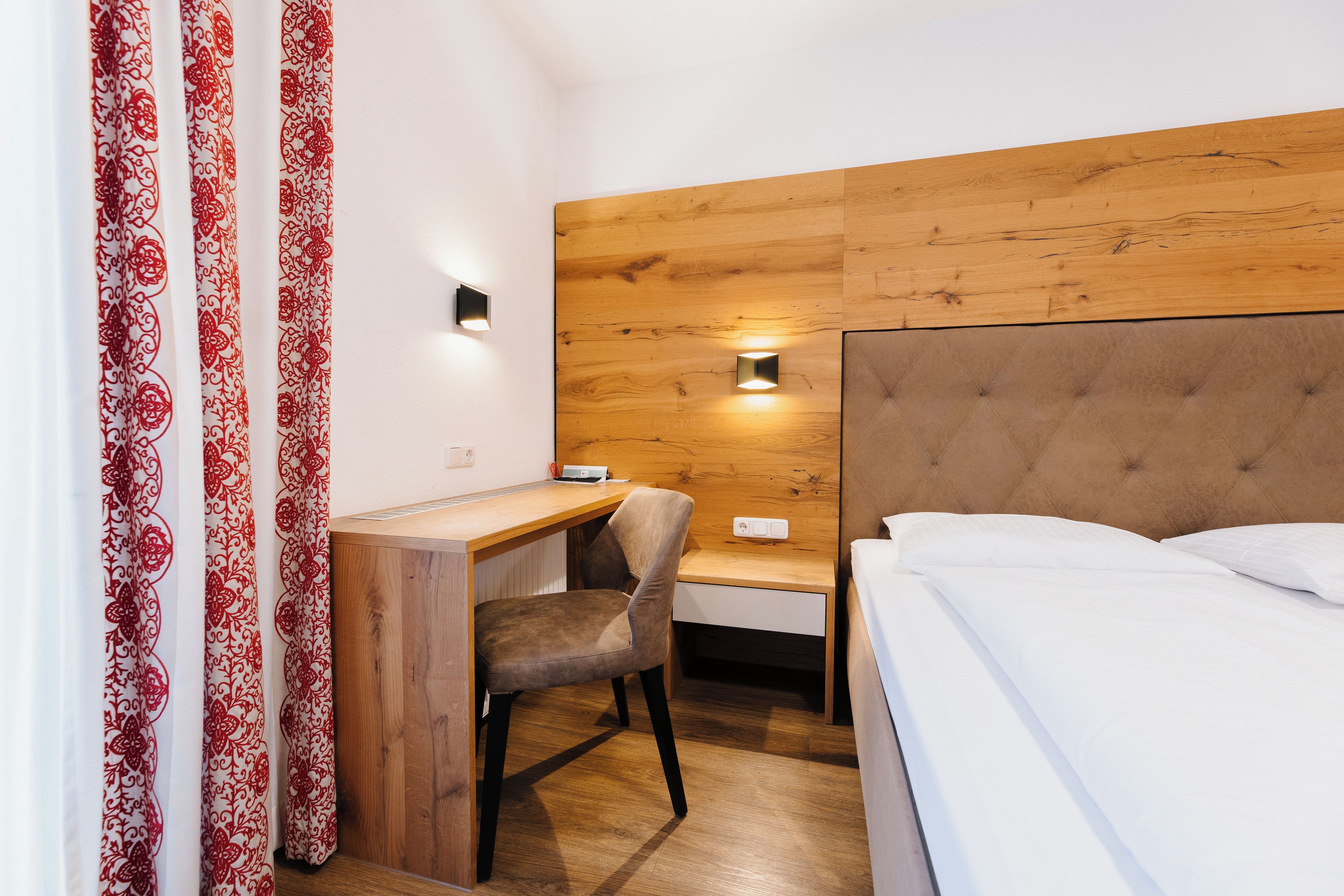 A hotel room with a wooden headboard, a desk with a chair, a bed with white bedding, and a wall-mounted light fixture.