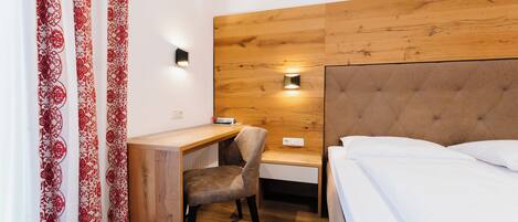 A hotel room with a wooden headboard, a desk with a chair, a bed with white bedding, and a wall-mounted light fixture.