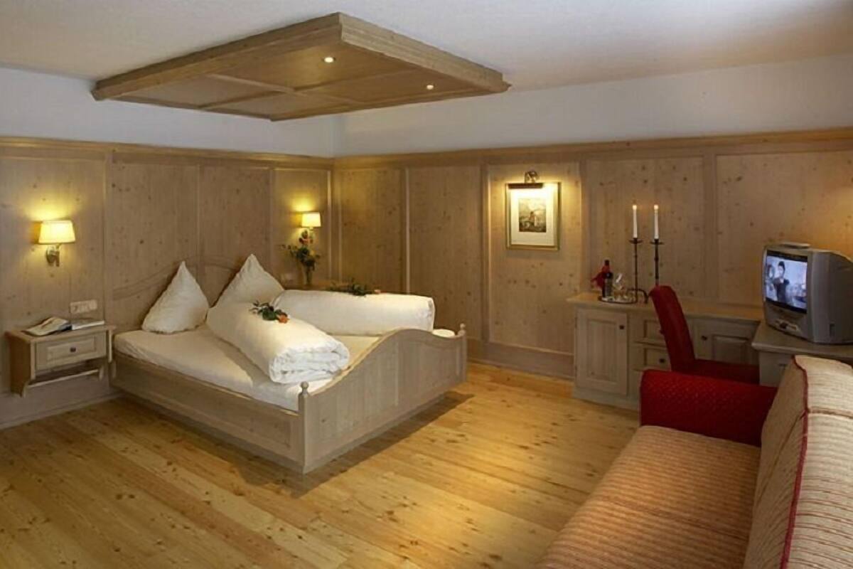 A bedroom with a wooden floor, a bed with white linens, a nightstand, a TV, and a red chair.