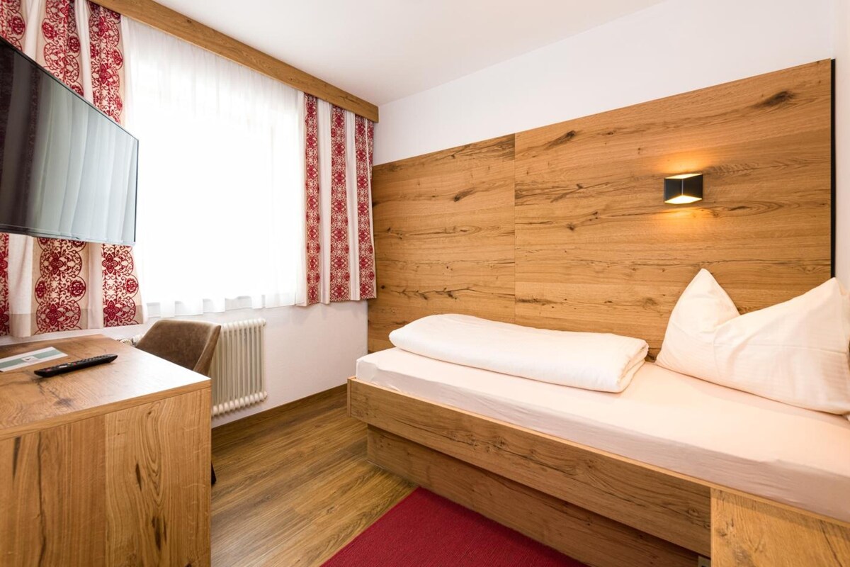 Een kleine hotelkamer met een houten hoofdeinde, een enkel bed, een bureau met stoel, een televisie en een raam met gordijnen met een patroon.