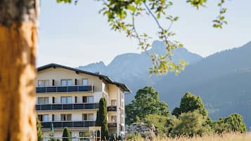 Ein mehrstöckiges Gebäude mit Balkonen, umgeben von einer Wiese und Bäumen, mit Bergen im Hintergrund.