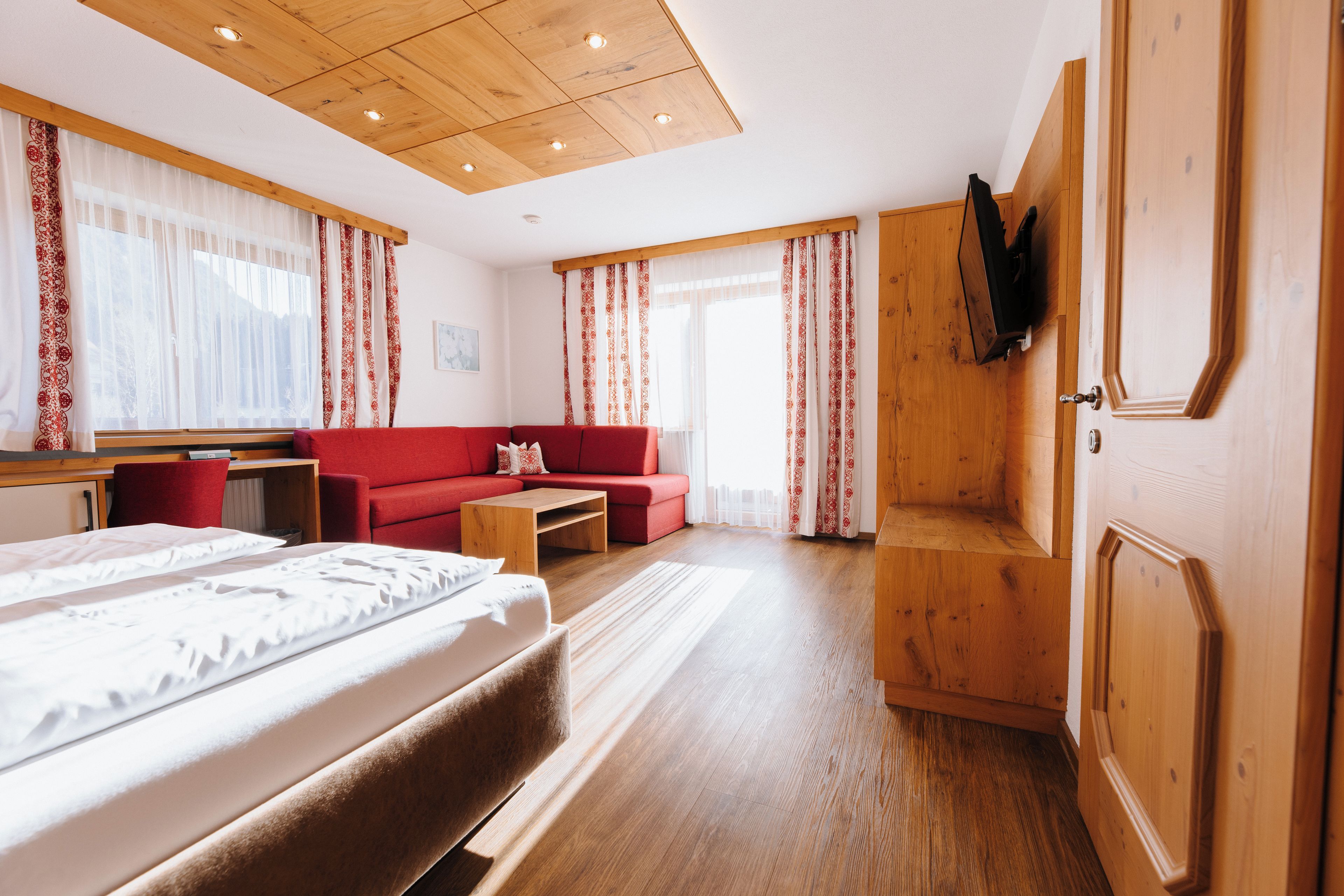 A hotel room with a bed, a red sofa, a wooden desk, and a TV mounted on the wall.