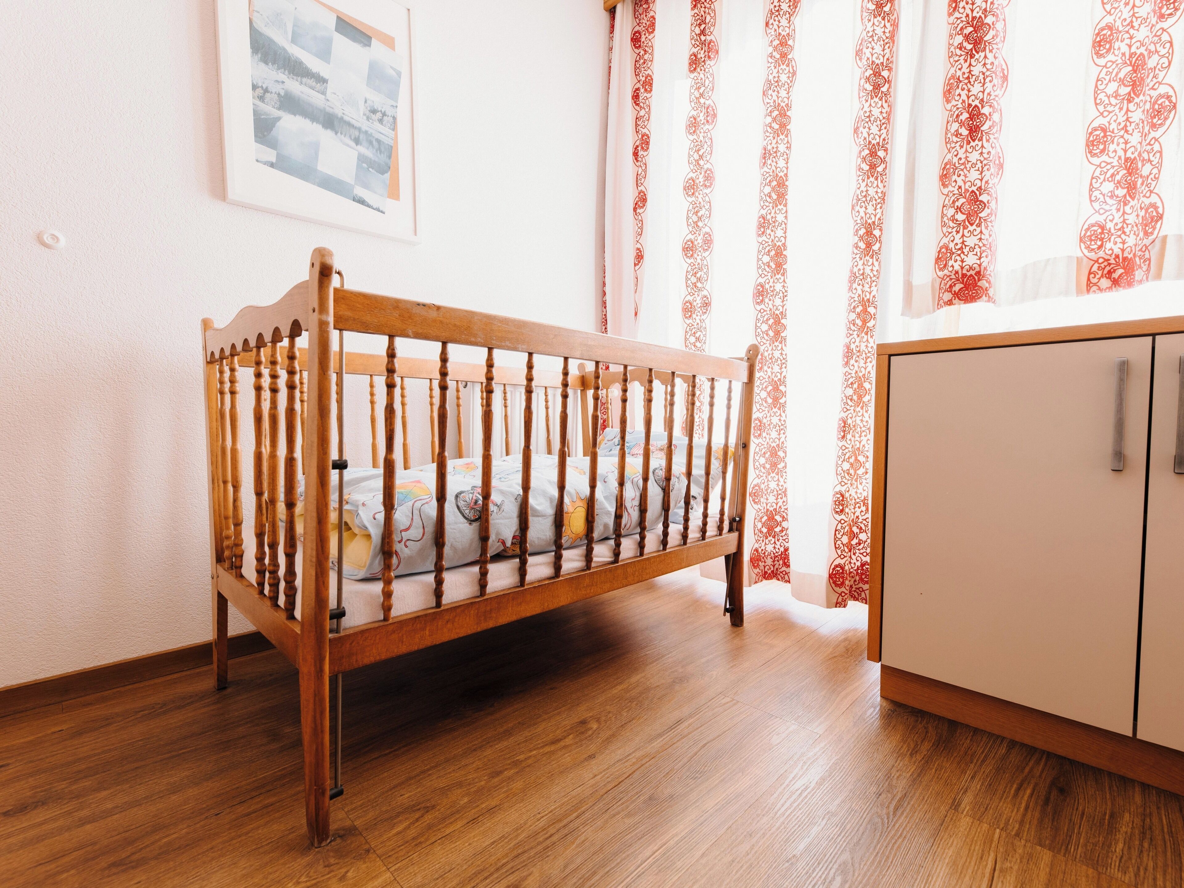 Ein hölzernes Kinderbett mit Matratze, eine weiße Kommode und ein gemusterter Vorhang.