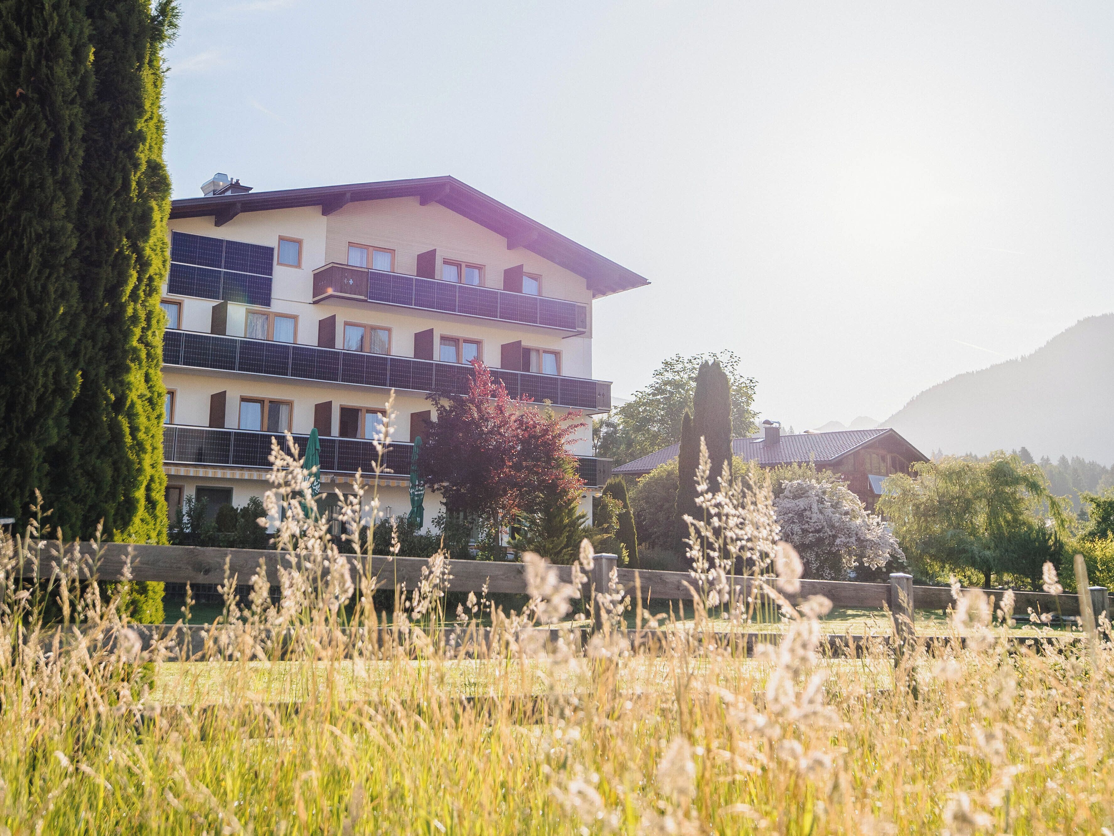 A multi-story building with balconies, surrounded by tall trees and a grassy field.