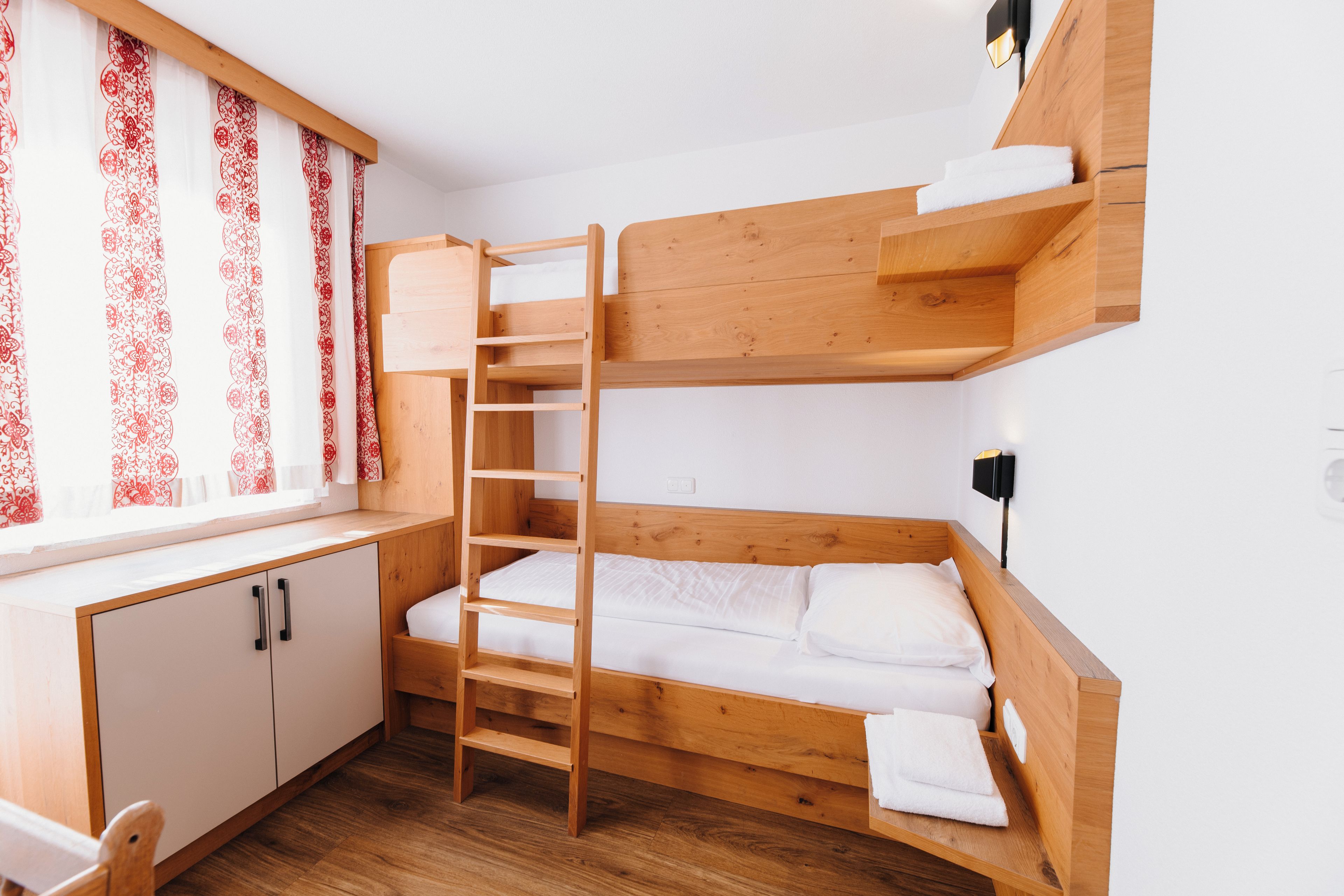 A bunk bed room with a wooden ladder, a desk with a chair, and a window with patterned curtains.