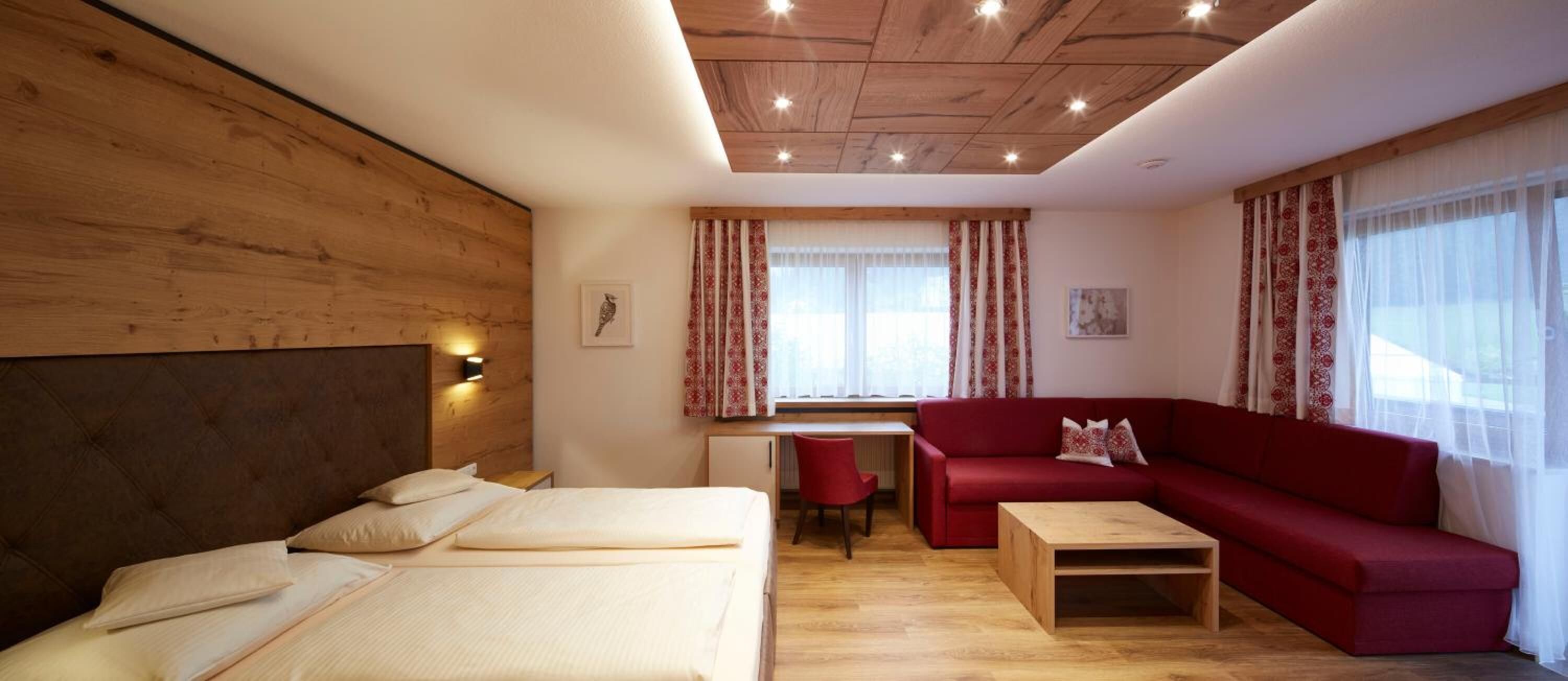 A hotel room with a wooden ceiling, a bed, a red sofa, a desk, and a window with curtains.
