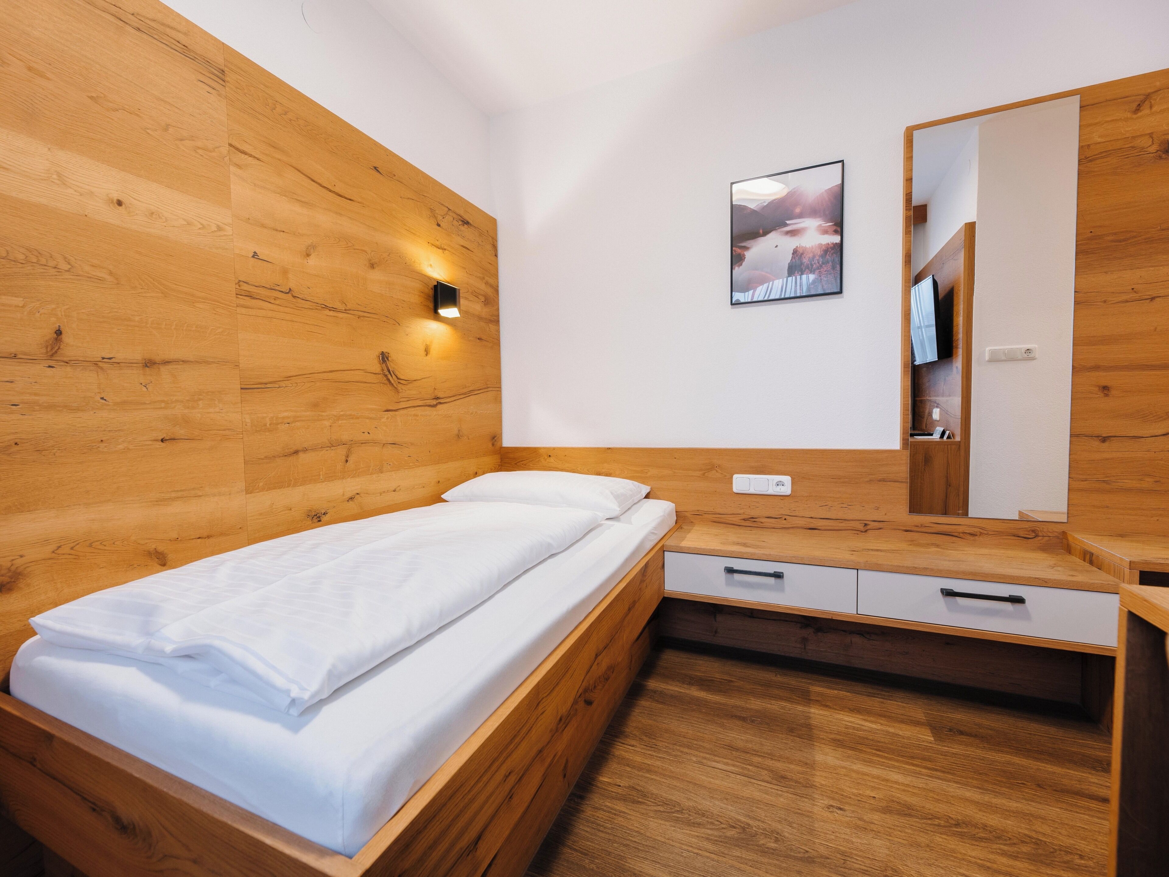 A bedroom with a wooden headboard, a bed with white bedding, a bedside table with drawers, a wall-mounted light, and a framed picture.