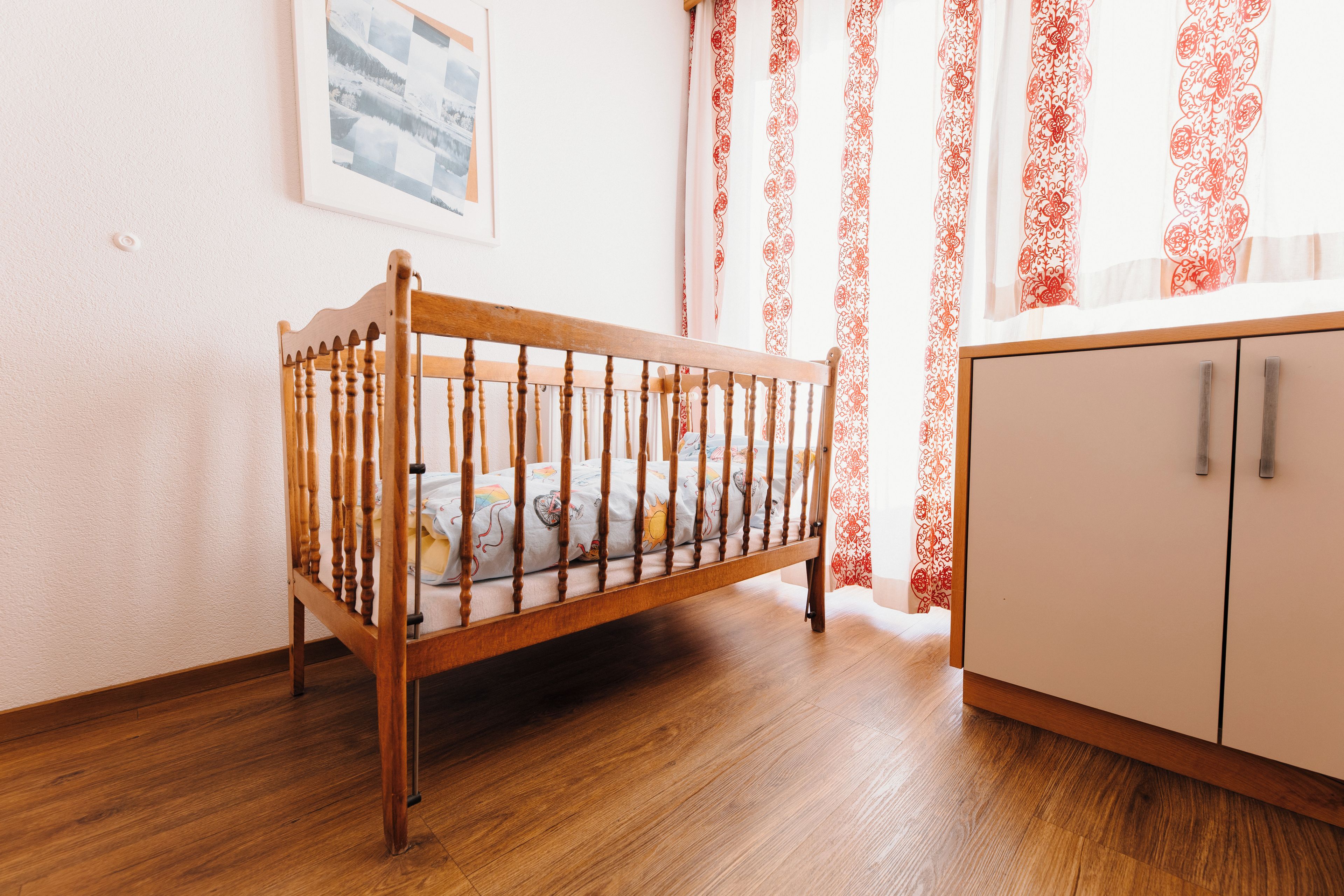 Ein hölzernes Kinderbett mit Matratze und Kissen, ein weißer Schrank mit zwei Schubladen sowie ein Fenster mit roten, gemusterten Vorhängen.