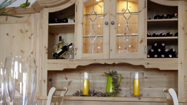 Una mesa de comedor con un frutero, copas de vino y un mueble de madera con botellas de vino.