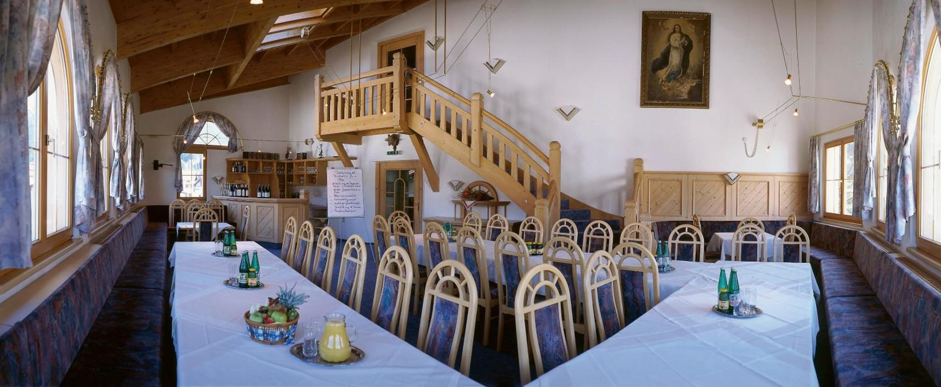 A dining area with long tables, wooden chairs, a staircase, and a mural on the wall.